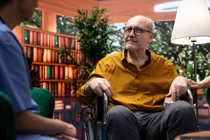 Senior woman comforting her husband with disability in a wheelchair while they talk to medical assistant, asking for professional advice