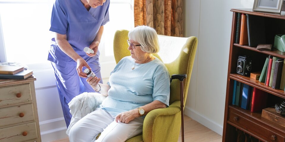 Senior woman in a chair receiving medication from a caregiver.,Caregiver assists a senior woman with medication.,Senior healthcare and medication concept.